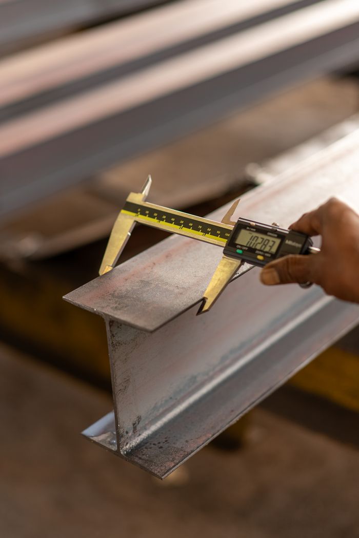 about-img Close-up of a caliper measuring a metal beam, showcasing precision engineering and tool usage.
