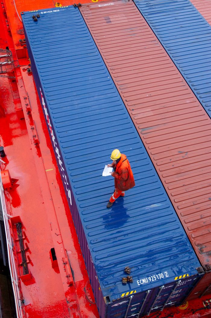 services-02 Overhead view of a worker inspecting containers on a ship deck in a maritime setting.